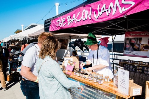A vendor in a green hat serves bacon jam to two customers at a market stall with a pink banner.