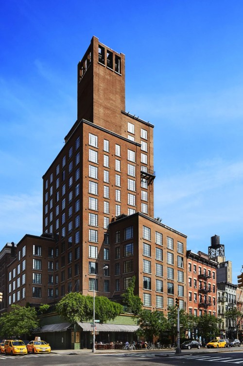A tall brick building with large windows and a clock tower, surrounded by trees and yellow taxis under a clear blue sky.