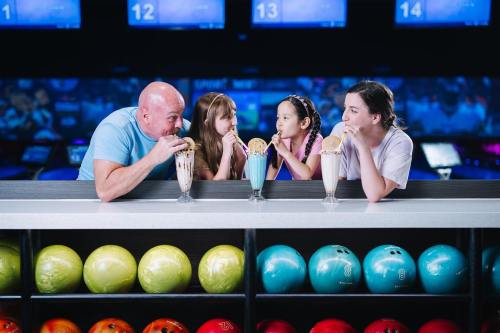 A man and three girls enjoy milkshakes at a bowling alley, with colorful bowling balls in the foreground.