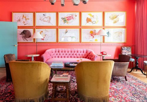 A vibrant lounge with a pink sofa, yellow chairs, and framed artwork on a coral wall, creating a cozy atmosphere.