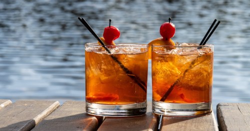 Two brandy old fashioned cocktails sit on a dock with a lake in the background.