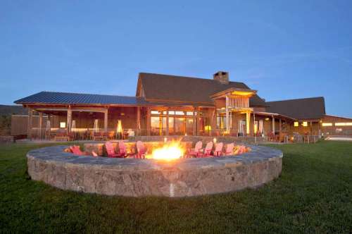 A cozy outdoor fire pit surrounded by chairs, with a modern lodge in the background under a clear blue sky.