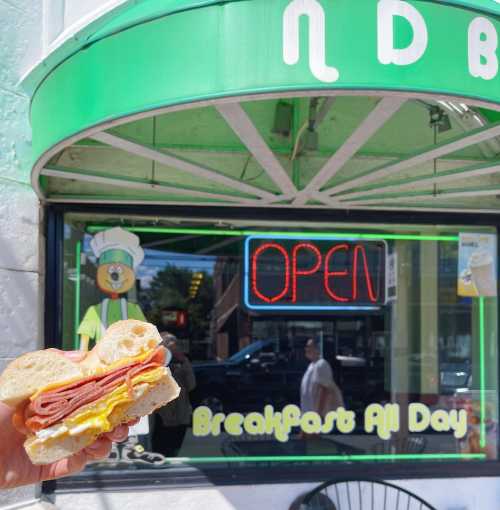 A hand holding a sandwich with deli meats and egg in front of a restaurant window displaying an "Open" sign.