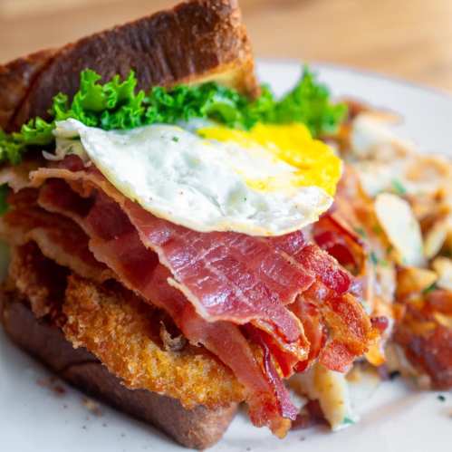 A hearty sandwich with layers of bacon, a fried egg, lettuce, and crispy bread, served with a side of hash browns.