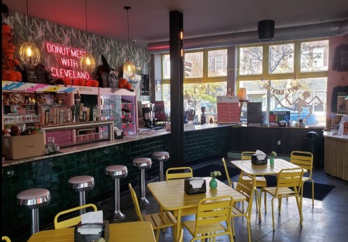 Bright café interior with yellow tables, green tiles, and a neon sign reading "DONUT MESS WITH CLEVELAND."