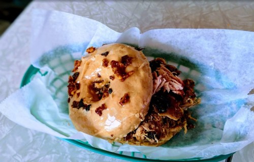 A close-up of a sandwich with pulled pork and a glazed bun, served in a green basket lined with white paper.