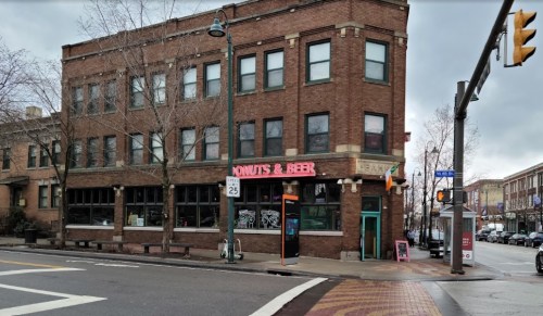 A brick building with a sign reading "Donuts & Beer" at the corner of a street, featuring large windows and a traffic light.