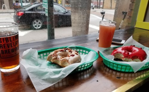 Two baskets with donuts—one with red icing and sprinkles, the other with white icing and bacon bits—beside drinks on a table.