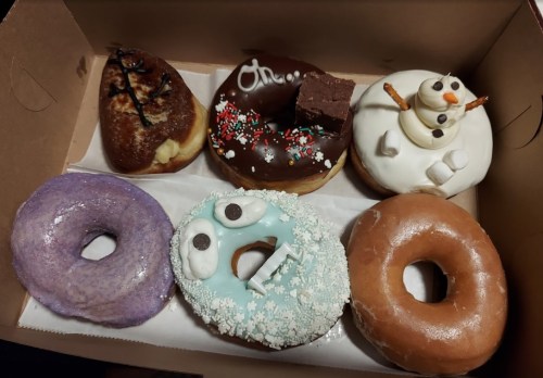 A box of six colorful donuts, including a snowman, a purple donut, and festive sprinkles.