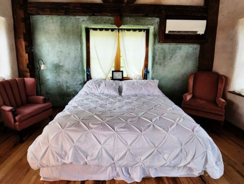 Cozy bedroom with a large bed, two armchairs, and a window with sheer curtains in a rustic setting.