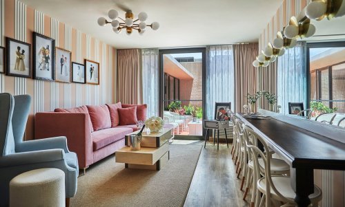 A stylish living room with a pink sofa, dining table, and large windows, featuring striped walls and modern decor.