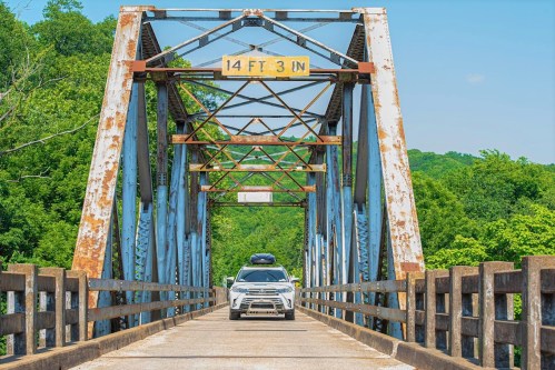 A white SUV crosses a weathered metal bridge surrounded by lush green trees under a clear blue sky.