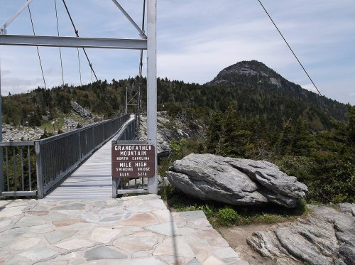 A suspension bridge leads to Grandfather Mountain, with rocky terrain and a peak in the background under a cloudy sky.