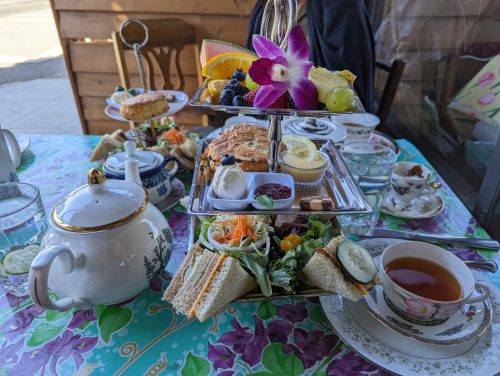 A beautifully arranged afternoon tea set with sandwiches, pastries, fruit, and a teapot on a floral tablecloth.