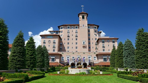 Historic hotel with a tall tower, surrounded by lush greenery and colorful flower beds under a clear blue sky.