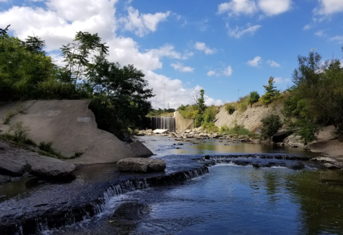A serene river scene with a small waterfall, rocky banks, and lush greenery under a partly cloudy sky.