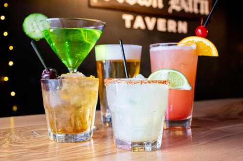A variety of colorful cocktails on a wooden table, including a martini, beer, and fruity drinks with garnishes.