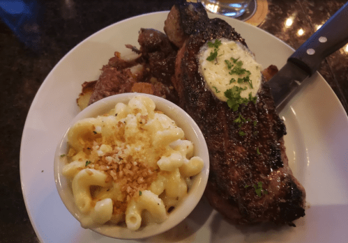 A plate featuring a grilled steak with herb butter, roasted potatoes, and a side of creamy macaroni and cheese.