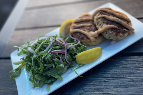 Two grilled patties drizzled with sauce, served with a side of arugula salad and lemon wedges on a white plate.