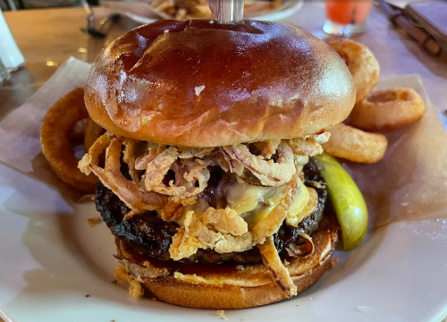 A stacked burger with crispy onions, cheese, and pickles, served with onion rings on the side.