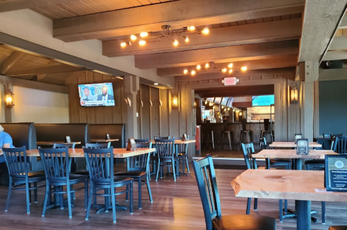 Interior of a restaurant featuring wooden tables, black chairs, and a TV on the wall, with a warm, inviting atmosphere.