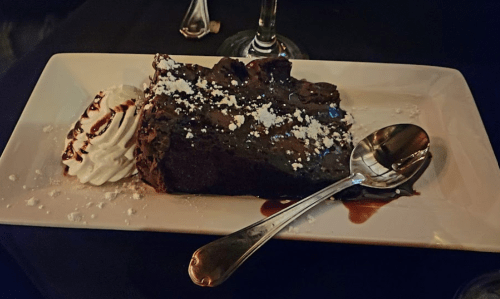 A slice of rich chocolate cake topped with powdered sugar, served with whipped cream and a drizzle of chocolate sauce.