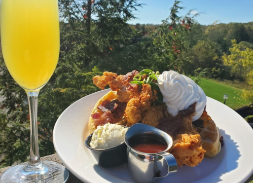 A plate of chicken and waffles topped with whipped cream, served with syrup and a mimosa in a glass.