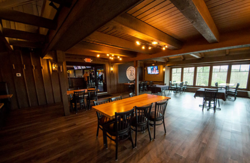 Interior of a rustic restaurant with wooden beams, tables, and large windows overlooking a natural view.