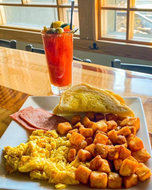 A plate of scrambled eggs, ham, golden potatoes, and toast, with a Bloody Mary cocktail in the background.