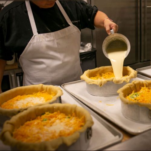 A chef pours liquid into pie crusts filled with cheese and vegetables, preparing them for baking.