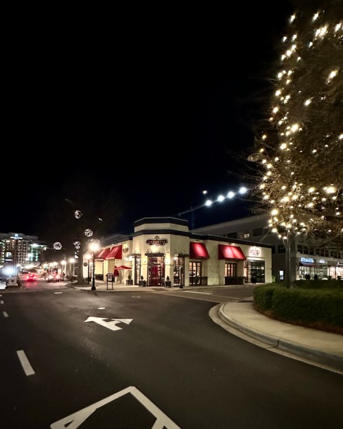 A street view at night featuring a restaurant with red awnings and festive lights on nearby trees.