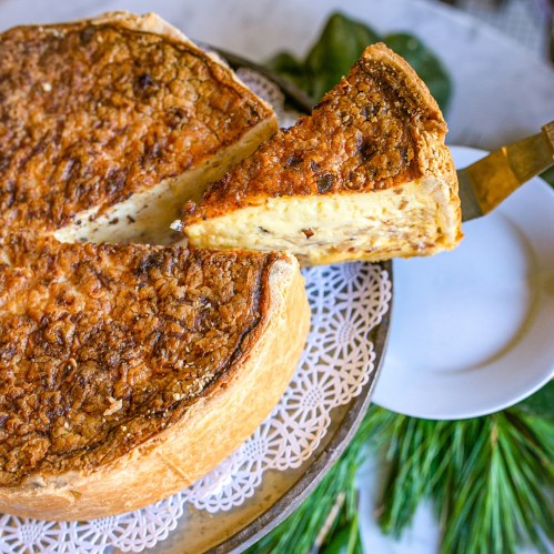 A round, golden-brown cheese pie with a slice being lifted, set on a decorative plate with greenery in the background.