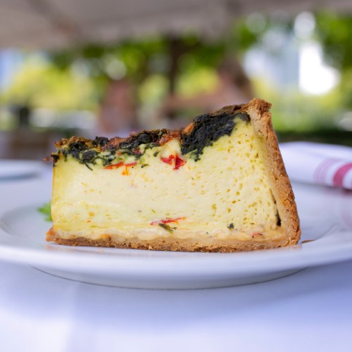 A slice of quiche with a golden crust, filled with spinach, red peppers, and a creamy yellow filling, served on a white plate.
