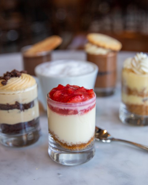 A variety of dessert shots, including cheesecake with strawberry topping, chocolate mousse, and whipped cream, on a marble surface.