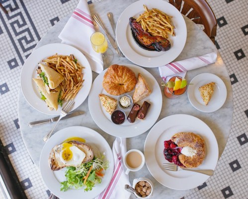 A table set with various dishes: fries, sandwiches, pastries, a salad, and a dessert, all on white plates.