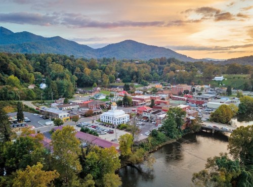 Aerial view of a small town by a river, surrounded by mountains, with colorful buildings and trees at sunset.