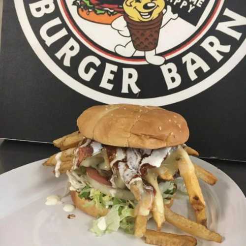 A large burger topped with bacon, lettuce, and sauce, served with fries, on a plate in front of a Burger Barn sign.