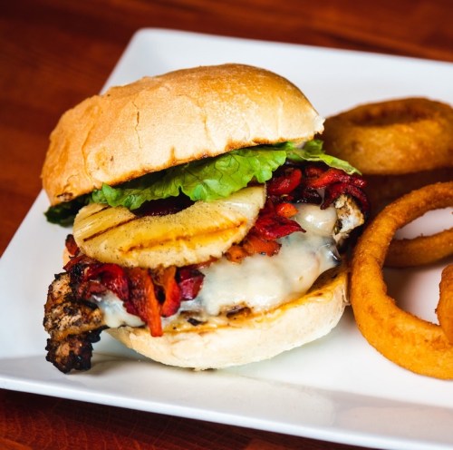 A grilled chicken burger topped with lettuce, pineapple, and red peppers, served with onion rings on a white plate.