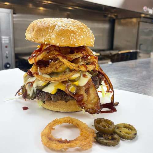 A towering burger with bacon, jalapeños, onion rings, and sauces on a sesame seed bun, served with an onion ring.