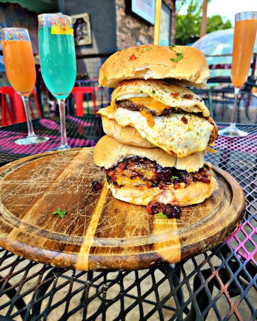 A towering burger with two fried eggs, bacon, and sauce, served on a wooden board with colorful cocktails in the background.
