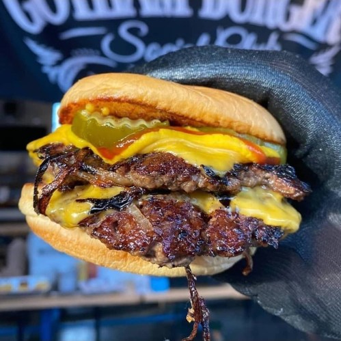 A hand holding a juicy burger with two beef patties, melted cheese, pickles, mustard, and ketchup on a soft bun.