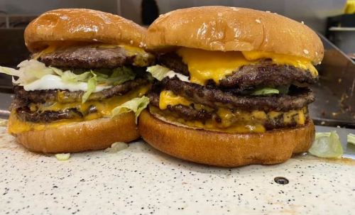 Two stacked burgers with multiple beef patties, melted cheese, lettuce, and sauces on sesame seed buns.