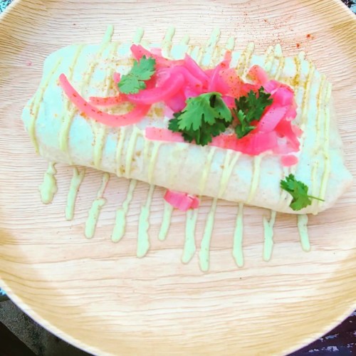 A burrito topped with pickled onions and cilantro, drizzled with sauce, served on a wooden plate.