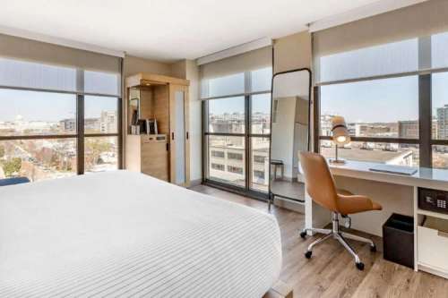 Modern hotel room with large windows, a desk, chair, and a mirror, offering a city view. Bright and spacious interior.
