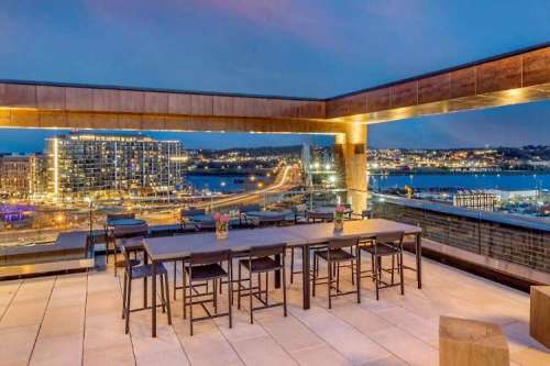 Rooftop terrace with a dining table overlooking a city skyline at dusk, featuring lights and a river view.
