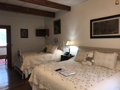 Cozy bedroom with two beds, decorative quilts, a lamp, and artwork on the walls, featuring wooden beams and warm lighting.