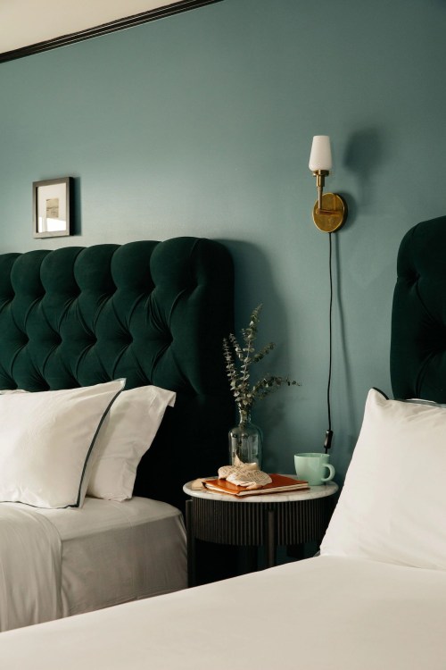 A cozy bedroom with two green tufted headboards, a side table with a plant, a cup, and a wall sconce.