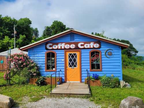 A bright blue coffee cafe with a wooden door, surrounded by greenery and flowers under a cloudy sky.