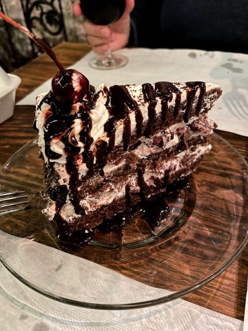 A slice of chocolate cake topped with whipped cream, cherries, and chocolate drizzle on a glass plate.