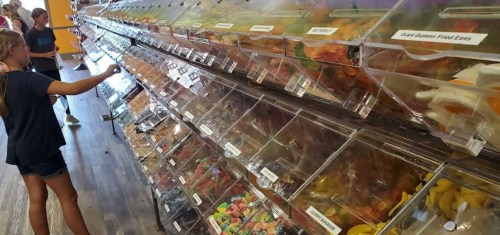 A colorful candy wall with various bins, while a child selects treats in a bright, cheerful shop.